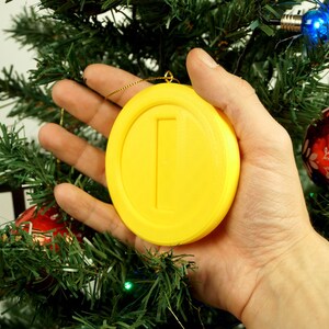Puede incluir: Un adorno de moneda amarillo impreso en 3D con una sola l&iacute;nea vertical en el centro, colgado de un &aacute;rbol de Navidad.