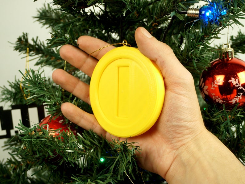 Puede incluir: Un adorno de moneda amarillo impreso en 3D con una sola l&iacute;nea vertical en el centro, colgado de un &aacute;rbol de Navidad.