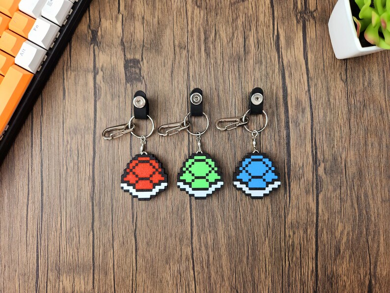 May include: Three keychains featuring pixelated turtle shells in red, green, and blue. Each keychain has a silver ring and a black clip.
