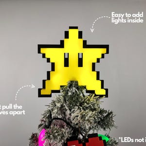 May include: A yellow pixelated star with a black outline, resembling a Super Mario Bros. power-up, is shown on top of a Christmas tree. The star is designed to be a Christmas tree topper and can be opened to add lights inside. The text "Easy to add lights inside" and "Just pull the halves apart" are visible in the image. The text "*LEDs not included" is also visible.