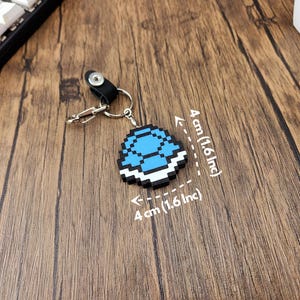 May include: A blue and white pixelated keychain shaped like a turtle shell. The keychain is 4 inches wide and 4 inches tall.