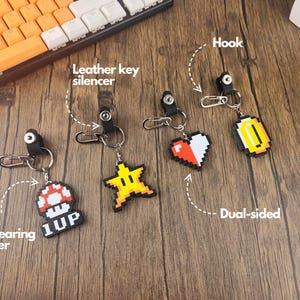 May include: Four different keychains with pixelated designs. The keychains feature a red heart, a gold coin, a yellow star, and a red mushroom with the text "1UP". Each keychain has a black leather key silencer and a silver hook.