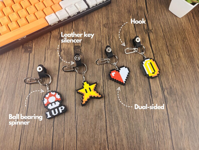 May include: Four different keychains with pixelated designs. The keychains feature a red heart, a gold coin, a yellow star, and a red mushroom with the text "1UP". Each keychain has a black leather key silencer and a silver hook.