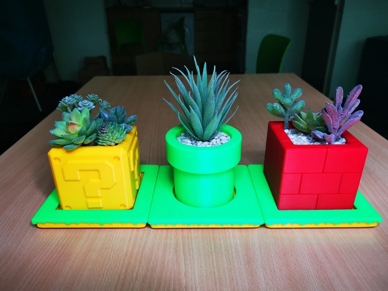 May include: Three small potted plants in green, yellow, and red planters shaped like a question mark block, a pipe, and a brick block from the Super Mario Bros. video game.