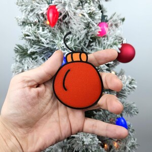 May include: A hand holding a red felt Christmas ornament with a black outline and a small orange loop at the top.