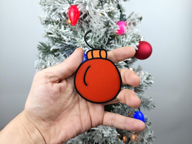 May include: A hand holding a red felt Christmas ornament with a black outline and a small orange loop at the top.