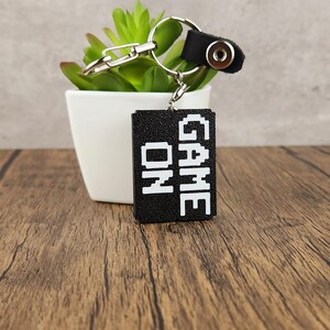 May include: Black keychain with a white 'GAME ON' inscription. The keychain is attached to a silver key ring and a black leather strap with a silver button.