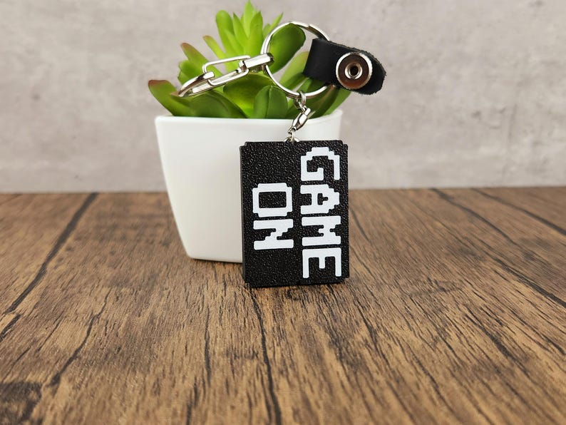 May include: Black keychain with a white 'GAME ON' inscription. The keychain is attached to a silver key ring and a black leather strap with a silver button.