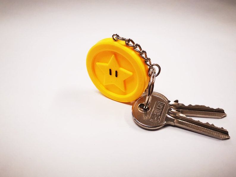 May include: A yellow star-shaped keychain with a black outline and two black dots for eyes. The keychain is attached to two silver keys.