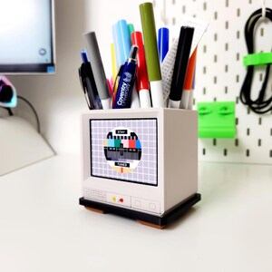 Pu&ograve; includere: Un organizzatore da scrivania bianco a forma di televisore con un motivo di test a barre di colore sullo schermo. L'organizzatore &egrave; pieno di varie penne e pennarelli, tra cui una penna blu con la scritta "Coventry University" stampata su di essa.