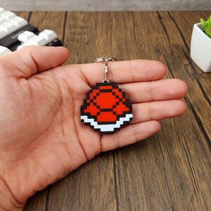 May include: A red and white pixelated Super Mario Bros. Koopa Shell keychain. The keychain is made of wood and has a silver metal ring.