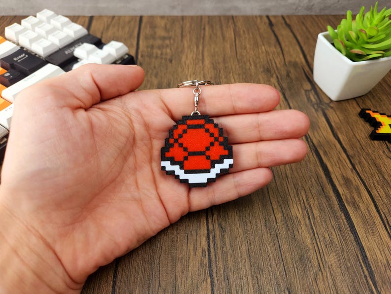 May include: A red and white pixelated Super Mario Bros. Koopa Shell keychain. The keychain is made of wood and has a silver metal ring.