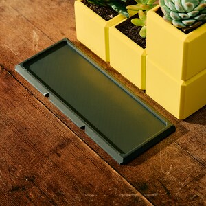 May include: A dark gray rectangular tray with rounded edges sits on a wooden surface.  Three yellow square planters with succulents are stacked on the right side of the tray.