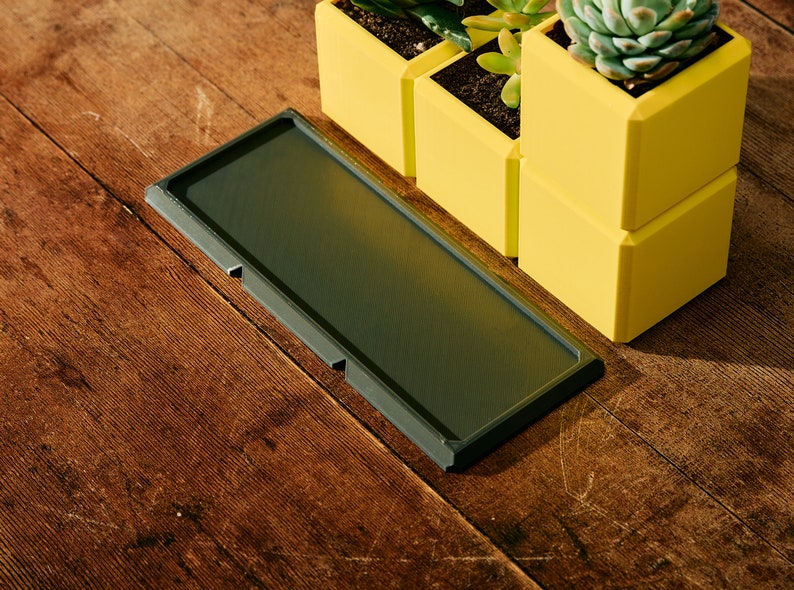 May include: A dark gray rectangular tray with rounded edges sits on a wooden surface.  Three yellow square planters with succulents are stacked on the right side of the tray.