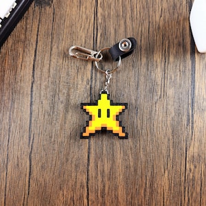 May include: A yellow pixelated star keychain with a black outline and a black ring. The keychain is hanging from a silver chain with a black leather loop.