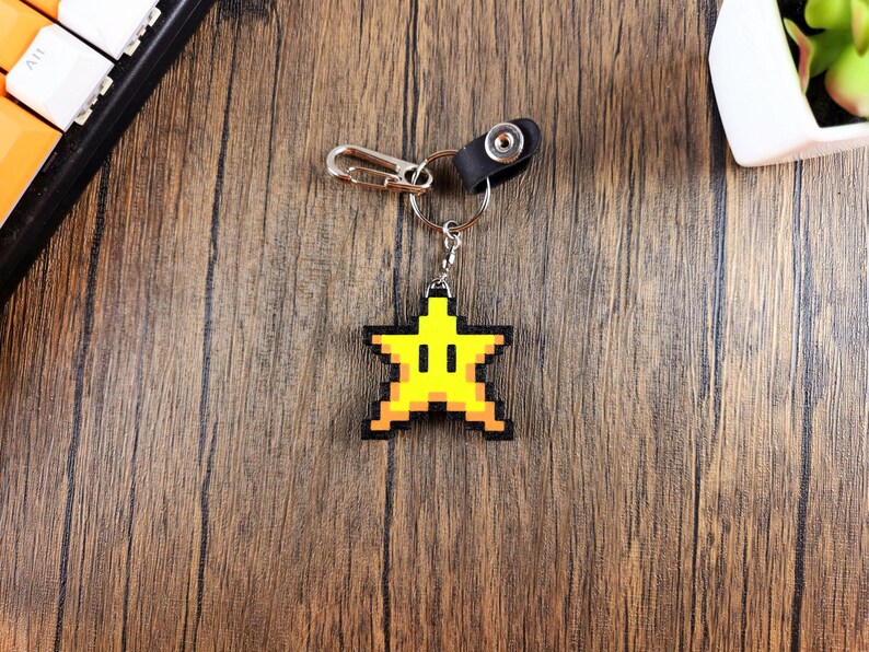 May include: A yellow pixelated star keychain with a black outline and a black ring. The keychain is hanging from a silver chain with a black leather loop.