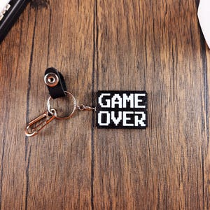 May include: A black keychain with a white pixelated "GAME OVER" design. The keychain is attached to a silver key ring and a black leather strap.