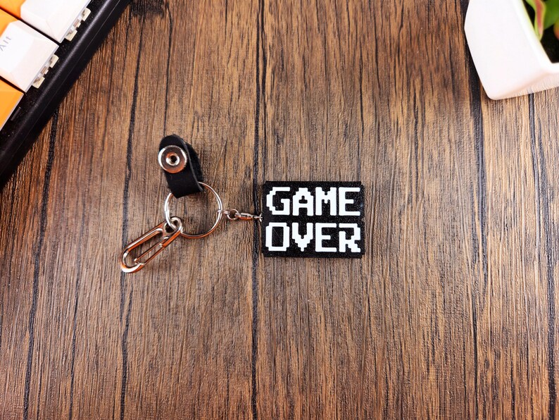 May include: A black keychain with a white pixelated "GAME OVER" design. The keychain is attached to a silver key ring and a black leather strap.