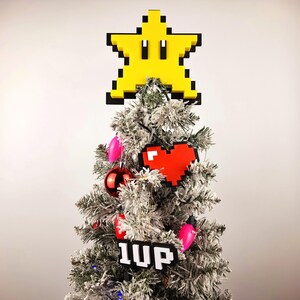May include: A snowy artificial Christmas tree decorated with a yellow star-shaped ornament, a red pixelated heart, and a black and white pixelated ornament that says "1UP".