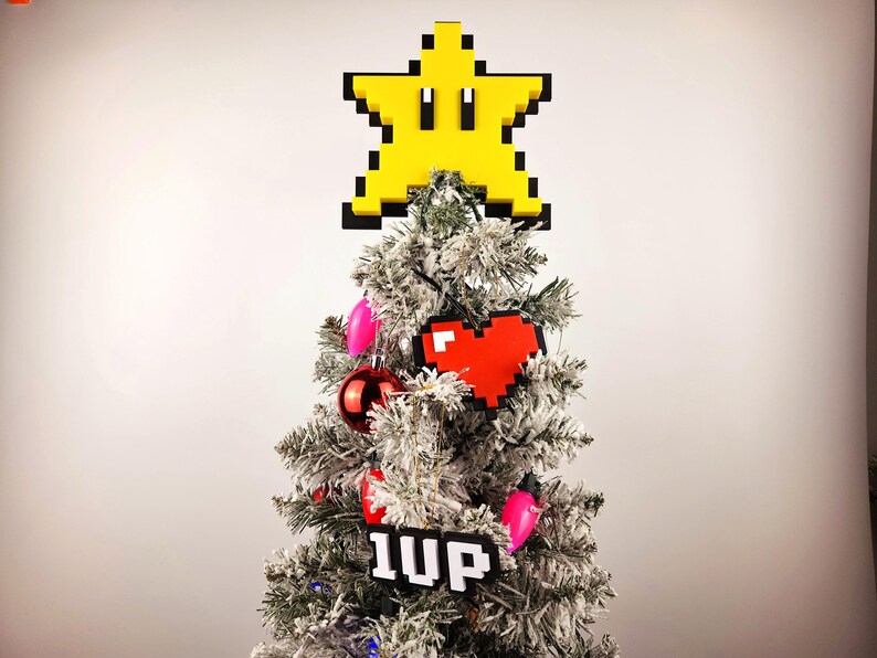 May include: A snowy artificial Christmas tree decorated with a yellow star-shaped ornament, a red pixelated heart, and a black and white pixelated ornament that says "1UP".
