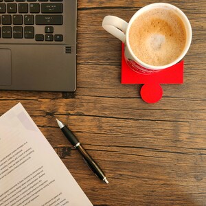 May include: A wooden desk with a laptop, a cup of coffee on a red puzzle-shaped coaster, a pen, and a document with text about a tangible sense of utility and enjoyment.
