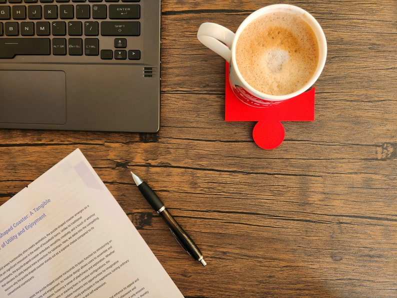 May include: A wooden desk with a laptop, a cup of coffee on a red puzzle-shaped coaster, a pen, and a document with text about a tangible sense of utility and enjoyment.
