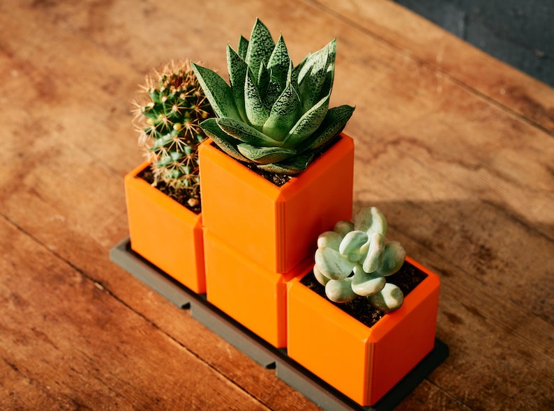 May include: Three orange square planters with different succulents. The planters are stacked on top of each other and are on a wooden surface.