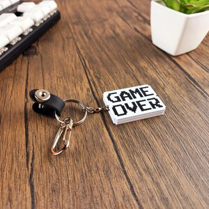 May include: A white keychain with a black pixelated "GAME OVER" design. The keychain is attached to a silver key ring and a black leather loop.