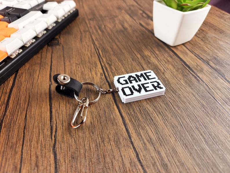 May include: A white keychain with a black pixelated "GAME OVER" design. The keychain is attached to a silver key ring and a black leather loop.
