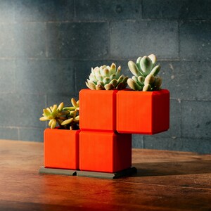 May include: A bright orange geometric planter, shaped like stacked blocks, contains three succulent plants. The planter rests on a dark grey base, with a dark brick wall behind. The wooden table provides a natural contrast.