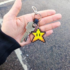 May include: A silver key chain with a yellow star-shaped charm. The star has a pixelated design and a smiling face.