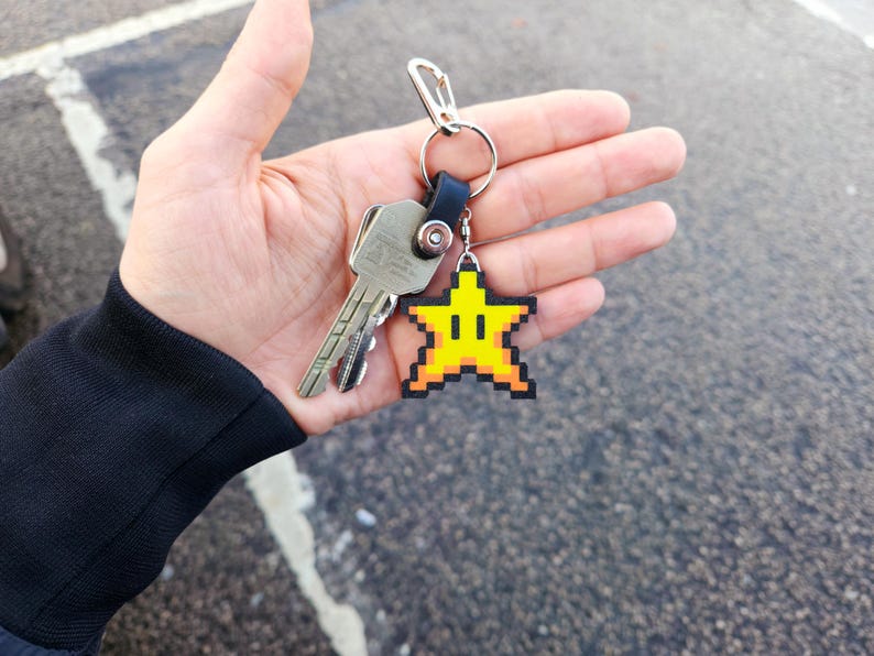 May include: A silver key chain with a yellow star-shaped charm. The star has a pixelated design and a smiling face.