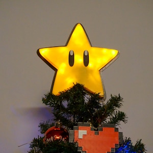 May include: A yellow star-shaped Christmas tree topper with black eyes and a light inside. The star is on top of a green Christmas tree with red and blue ornaments.