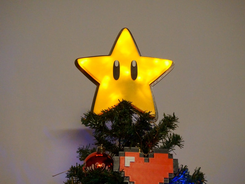 May include: A yellow star-shaped Christmas tree topper with black eyes and a light inside. The star is on top of a green Christmas tree with red and blue ornaments.