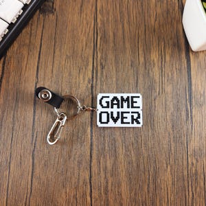 May include: A white keychain with a black pixelated "GAME OVER" design. The keychain has a silver metal ring and a black leather strap with a silver metal clasp.