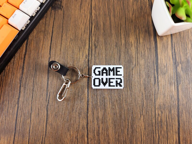 May include: A white keychain with a black pixelated "GAME OVER" design. The keychain has a silver metal ring and a black leather strap with a silver metal clasp.