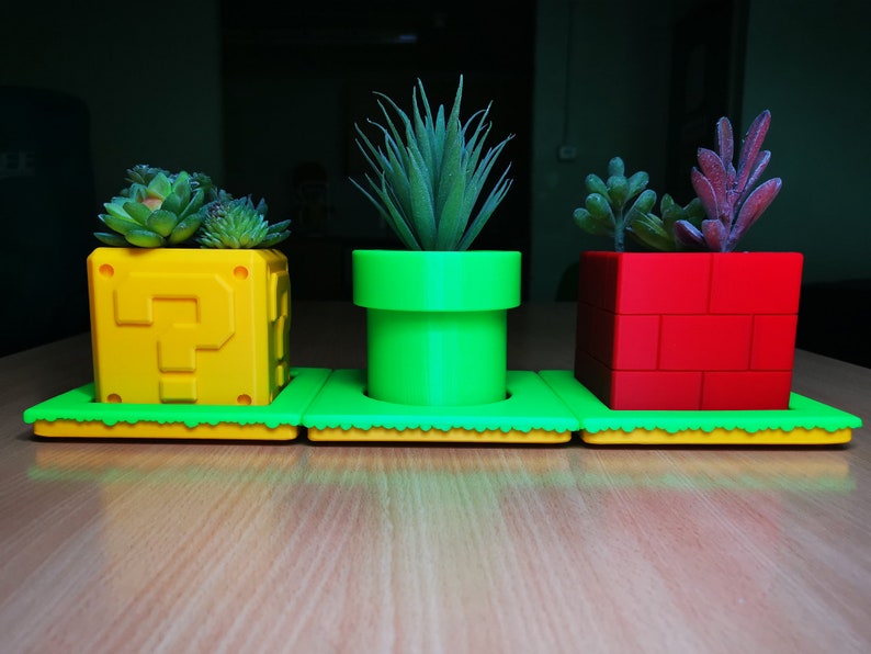 May include: Three colorful plant pots with green plants. The pots are shaped like a question mark block, a pipe, and a brick block, all from the Super Mario Bros. video game.