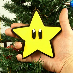 Puede incluir: Un adorno de Navidad de color amarillo en forma de estrella con contornos negros y dos grandes ojos negros. El adorno est&aacute; siendo sostenido por una mano frente a un &aacute;rbol de Navidad.