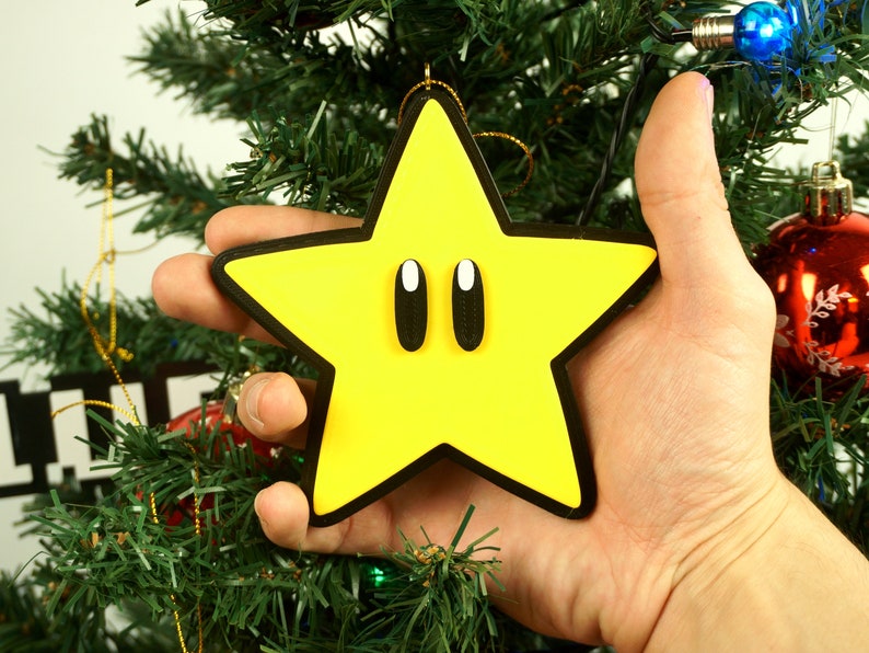 Puede incluir: Un adorno de Navidad de color amarillo en forma de estrella con contornos negros y dos grandes ojos negros. El adorno est&aacute; siendo sostenido por una mano frente a un &aacute;rbol de Navidad.