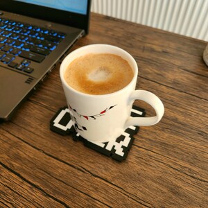 May include: A white ceramic mug with a festive design sits on a black and white pixelated coaster. The mug is filled with a light brown coffee beverage with a foam design on top.