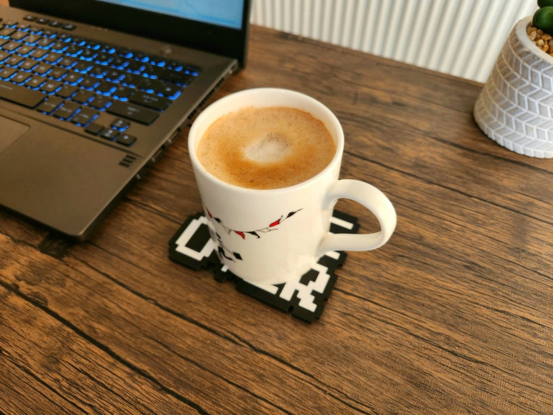 May include: A white ceramic mug with a festive design sits on a black and white pixelated coaster. The mug is filled with a light brown coffee beverage with a foam design on top.