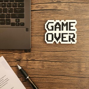 Puede incluir: Una pegatina blanca con texto pixelado negro que dice "GAME OVER" sobre una superficie de madera. Tambi&eacute;n se puede ver un port&aacute;til, un bol&iacute;grafo y una hoja de papel.