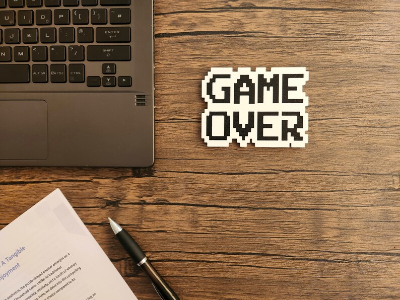 Puede incluir: Una pegatina blanca con texto pixelado negro que dice "GAME OVER" sobre una superficie de madera. Tambi&eacute;n se puede ver un port&aacute;til, un bol&iacute;grafo y una hoja de papel.