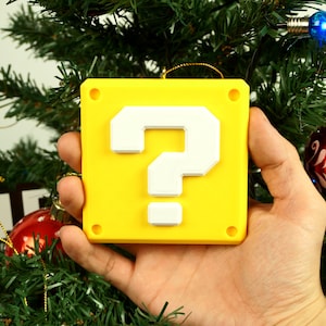 Puede incluir: Un adorno navide&ntilde;o de bloque de signo de interrogaci&oacute;n amarillo con un signo de interrogaci&oacute;n blanco. El adorno est&aacute; colgado de un &aacute;rbol de Navidad verde.