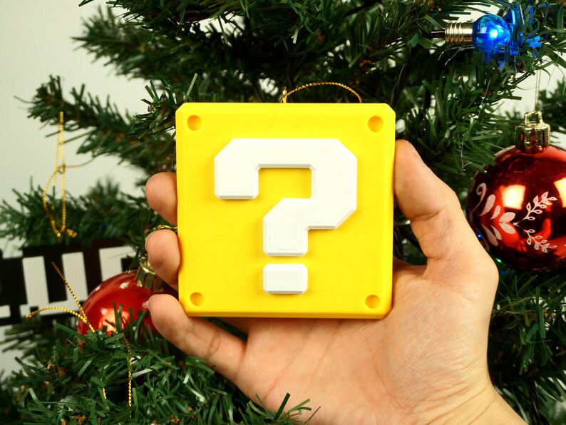 Puede incluir: Un adorno navide&ntilde;o de bloque de signo de interrogaci&oacute;n amarillo con un signo de interrogaci&oacute;n blanco. El adorno est&aacute; colgado de un &aacute;rbol de Navidad verde.
