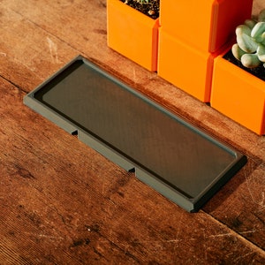 May include: A dark gray rectangular tray with rounded corners sits on a wooden surface.  The tray has three small raised sections on the bottom.  Three orange square planters with succulents are visible in the background.