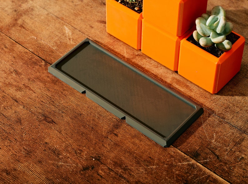 May include: A dark gray rectangular tray with rounded corners sits on a wooden surface.  The tray has three small raised sections on the bottom.  Three orange square planters with succulents are visible in the background.