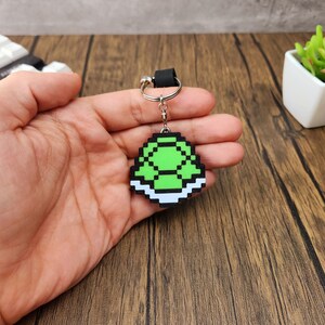 May include: A green and white pixelated turtle shell keychain. The shell is green with a white border and is attached to a silver key ring.