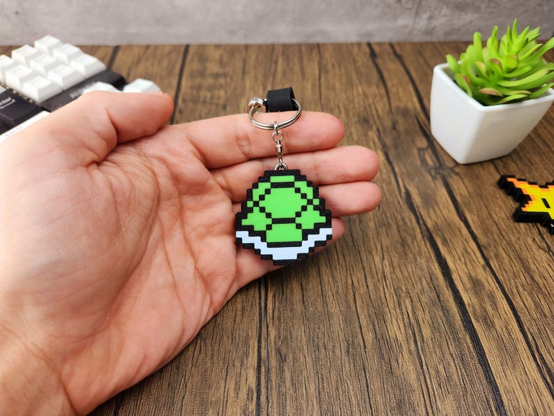 May include: A green and white pixelated turtle shell keychain. The shell is green with a white border and is attached to a silver key ring.