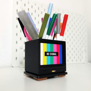 Pu&ograve; includere: Un organizzatore da scrivania nero a forma di televisore con uno schermo colorato "NESSUN SEGNALE" e una variet&agrave; di penne e pennarelli all'interno.